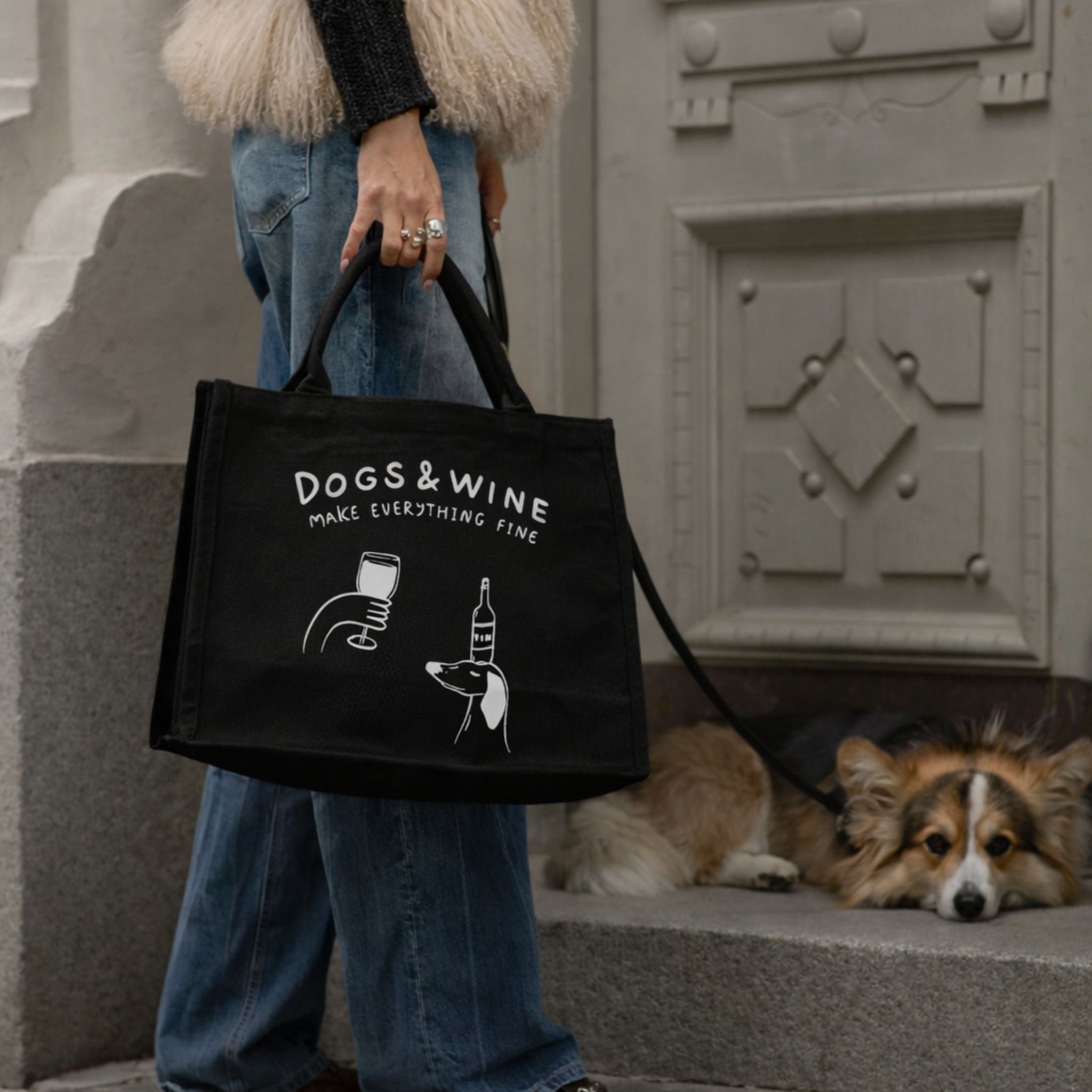 Person holding a black tote bag with 'DOGS & WINE make everything fine' text and a dog lying on steps in the background.