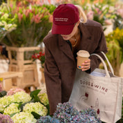 Person wearing a red cap and brown coat, holding a coffee cup and a tote bag with 'Dogs & Wine' branding, standing among flowers.