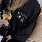Person wearing a black sweatshirt with 'Cats & Wine' text and graphics, holding a glass of white wine.